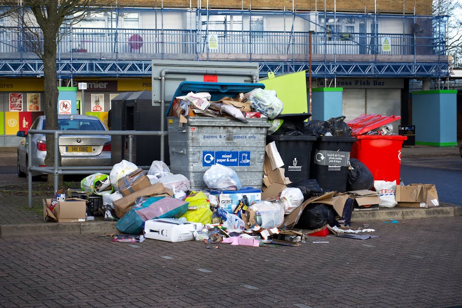 A large pile of mixed household rubbish and waste bags is overflowing from multiple black, red, and grey rubbish bins situated on a paved area in front of a commercial building with a blue scaffold structure. The waste includes cardboard boxes, paper, plastic bags, and miscellaneous packaging materials, with some items spilling onto the surrounding pavement and street. A grey suburban car is parked nearby, with a metal railing separating it from the rubbish. The background features storefronts with signs and windows, indicating a retail or commercial area. The scene appears to be an example of bulk rubbish accumulated outside a property, possibly awaiting collection or disposal, reflecting the informal disposal methods often seen in private waste handling scenarios. This visual relates to waste management and the importance of adhering to local rubbish rules or alternative collection services, like independent clearance, especially in urban settings like Crystal Palace.