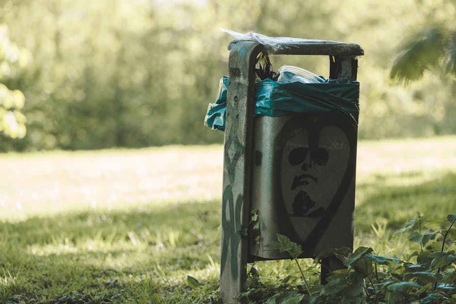 A weathered metal waste bin situated outdoors on a grassy area with a backdrop of blurred green foliage and sunlight filtering through trees. The bin features a graffiti illustration of a stylized face with large, dark eyes, a prominent nose, and an open mouth, painted in black. The top of the bin appears to be covered with a bent and slightly torn lid, with some blue and transparent plastic bags protruding from the open side, suggesting recent use for rubbish disposal. The bin's surface shows signs of rust and wear, with some greenish moss or algae growth on the side. Surrounding the bin are small leafy plants growing at its base. The scene indicates a location suitable for independent waste collection or private rubbish clearance, with the environment emphasizing the importance of proper waste disposal methods outside urban areas and the potential for local rubbish removal services like those offered by rubbishclearancecrystalpalace.org.uk to handle such waste management needs effectively.