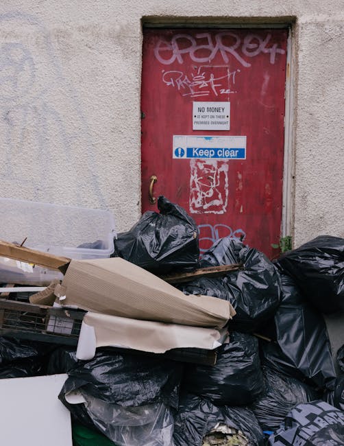 A collection of black rubbish bags and discarded packaging materials piled against a red metal door on an outdoor wall. The door is weathered with graffiti tags in white and pink spray paint, along with a blue sticker that reads 'Keep clear.' Above the sticker, there is a white sign stating 'NO MONEY' and warning about the consequences of illegal dumping. The rubbish bags appear full and are situated on a paved surface, with some items like flattened cardboard boxes and wooden pieces visible among the waste. The surrounding environment is part of an urban exterior, likely adjacent to a building or alleyway, illustrating a scene of private waste accumulation that could relate to alternative waste handling or on-site rubbish clearance services provided by companies like rubbishclearancecrystalpalace.org.uk.