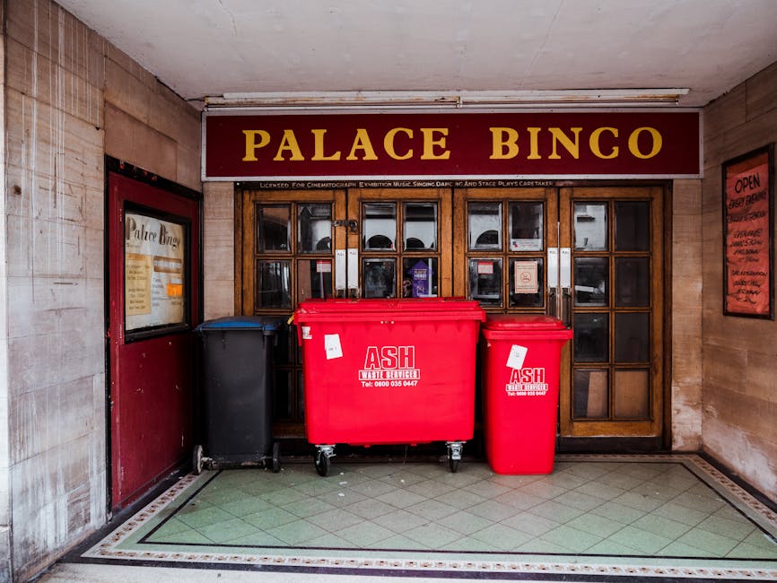 The image depicts the entrance of a venue named 'Palace Bingo' with a maroon sign above the wooden double doors featuring multiple glass panels. In front of the doors, there are three large wheeled bins for waste collection: a black bin on the left, a large red bin with white labels in the middle, and a smaller red bin with a white label on the right. The bins are positioned on a tiled floor with a green and cream pattern, adjacent to a beige wall with a framed poster on the left side and a red poster on the right. The environment appears to be an indoor or sheltered area, with lighting that highlights the bins and the wooden structure of the doors, indicative of private waste handling or alternative rubbish removal options supported by waste management services such as those provided by rubbishclearancecrystalpalace.org.uk.