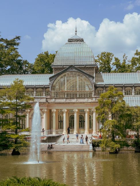 A historic glass and iron conservatory with a domed roof and ornate architectural details, situated within a park setting. The building features large arched windows and intricate decorative elements along its facade, with a prominent central entrance framed by classical columns. Surrounding the conservatory, there are tall, leafy trees with green foliage, partially obscuring the sides of the structure. In front, a reflecting pond extends across the foreground, with a tall water fountain erupting from its center, creating a vertical spray of water. Several people are visible on the wide stone steps leading up to the conservatory, some walking and others standing, enjoying the outdoor environment. The scene is lit by natural daylight, with a partly cloudy sky overhead, casting soft shadows and highlighting the building’s glass and metal finishes, characteristic of a historical botanical glasshouse that may be featured in parks or gardens. This setting emphasizes a serene outdoor space that can be associated with the importance of maintaining clean and accessible public areas, possibly supported by independent waste management services like those provided by rubbish clearance specialists.