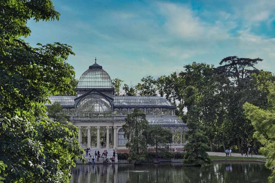 A large historic glass and stone conservatory situated in a park setting, with a symmetrical classical design featuring tall columns and a domed roof at its center. The building is surrounded by lush green trees and shrubs, some partially obscuring the structure. In the foreground, a pond reflects the building and the surrounding natural landscape, with a fountain visible near the center of the pond. The sky above is bright and partly cloudy, providing natural light that enhances the greenery and the architectural details of the conservatory. The scene is tranquil, with a few pigeons on the grassy area in the front, indicating a peaceful outdoor environment. This image, associated with rubbish clearance services, highlights the natural environment that benefits from efficient waste management and on-site clearance solutions provided by companies operating in the area, supporting clean and preserved outdoor spaces near prominent landmarks.