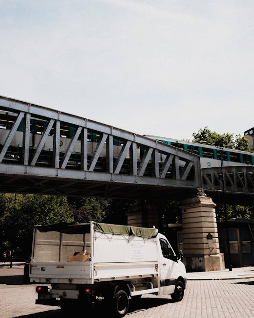 A white flatbed commercial vehicle with an open cargo area is parked on a paved surface beneath a large railway viaduct. The vehicle's cargo area is partially covered with a green tarpaulin, secured with ropes, and appears to contain some cardboard boxes or debris, suggesting a waste collection activity. The viaduct overhead is constructed from metal trusses painted in shades of gray, supported by large, rounded stone pillars. The scene is set in an urban environment with a background of trees and a clear sky, indicative of daytime lighting. The location implies a setting where private or independent waste removal services, such as rubbish clearance, might operate, especially with the visible debris and equipment suitable for on-site rubbish collection, which complements or offers alternatives to local authority disposal. The overall scene reflects a typical rubbish removal operation related to private waste handling, with the presence of the vehicle implying readiness for rubbish collection or transportation in a Crystal Palace area context.